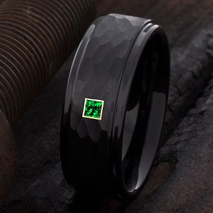 Op de afbeelding: Een zwarte ring met een gehamerde textuur en een vierkante, smaragdgroene edelsteen gezet in een gouden frame. De ring heeft een gepolijste, gladde binnenband. De ring is een sieraad.