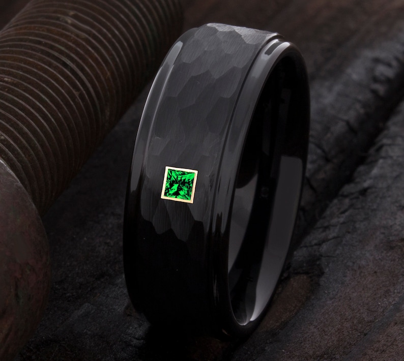 Op de afbeelding: Een zwarte ring met een gehamerde textuur en een vierkante, smaragdgroene edelsteen gezet in een gouden frame. De ring heeft een gepolijste, gladde binnenband. De ring is een sieraad.