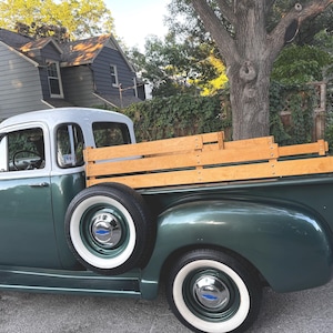 May include: Vintage green pickup truck with a white roof and wooden bed. The truck has white-walled tires and chrome hubcaps. The truck is parked on a paved road in front of a house.