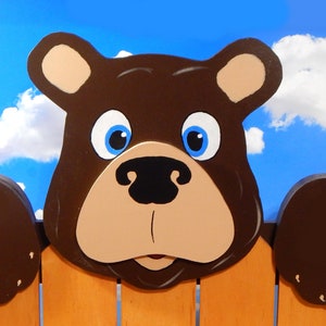 May include: A brown wooden bear with blue eyes and a black nose peeking over a wooden fence. The bear has brown fur and a tan face. The fence is brown and has white pickets.