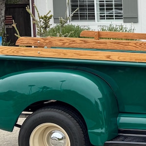 May include: Close-up of a vintage green pickup truck. The truck bed features light-colored wood paneling. The tires have cream-colored rims. The truck's body is a glossy, dark green color.