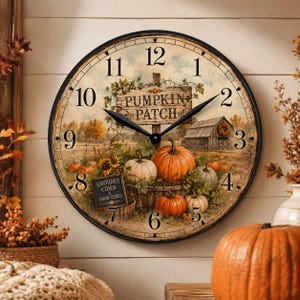 May include: A round wall clock with a black frame featuring a printed scene of a pumpkin patch, a wooden sign reading PUMPKIN PATCH, a barn, and a small sign reading HAYRIDES CIDER & GOOD TIMES. The clock face has black hands and black numerals.