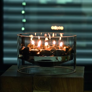 May include: A clear glass tabletop fire pit with a metal base and black stones. The fire pit is lit with several small flames.