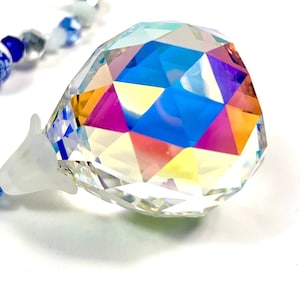 May include: A faceted crystal prism with a teardrop shape, reflecting a spectrum of colors. A string of blue and white beads is visible in the background. The prism is suspended by a clear, plastic-like attachment.