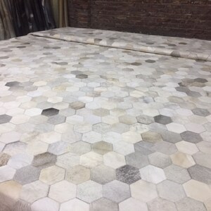 May include: A large rug with a geometric pattern of hexagonal shapes in neutral tones. The rug features a mix of white, grey, and tan hues, creating a textured, natural look. The rug is likely made of leather or a similar material.