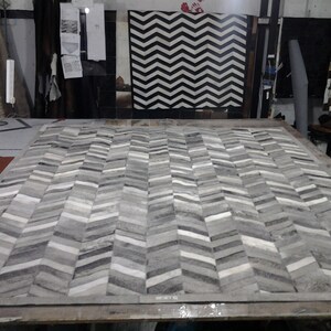 May include: A gray and white chevron patterned rug made from cowhide. The rug is made up of individual pieces of cowhide sewn together to create the chevron pattern.