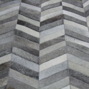 May include: A gray and white chevron patterned rug made from cowhide. The rug is made up of many different pieces of cowhide, each with a different shade of gray or white. The rug is sewn together with a white thread.