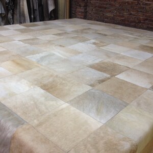 Puede incluir: Una alfombra grande, blanca, de patchwork hecha de piel de vaca. La alfombra está hecha de cuadrados de diferentes tamaños y tonos de blanco y beige.
