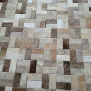 May include: A patchwork rug made of cowhide in shades of brown, beige, and white. The rug is made up of many different sized rectangular pieces of leather, sewn together to create a unique and interesting pattern.