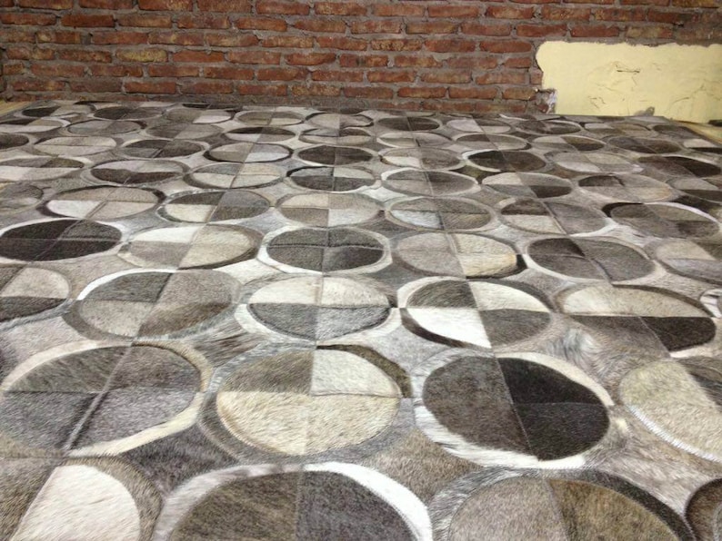 May include: A close-up of a gray and white patterned rug made of cowhide. The rug is made up of many different shapes and sizes of cowhide patches, creating a unique and interesting design.