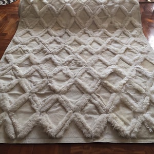 Puede incluir: Una alfombra blanca y esponjosa con un patrón de diamantes. La alfombra está hecha de un material suave y mullido y tiene un brillo sutil.