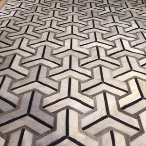 May include: Geometric patterned area rug in shades of white, gray, and black. The rug features a repeating three-pronged design. The rug is rectangular and appears to be made of a soft material, suitable for a living room or bedroom.