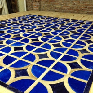 May include: A geometric patterned rug made of leather in shades of blue, tan, and black. The rug features a repeating pattern of circles and squares.