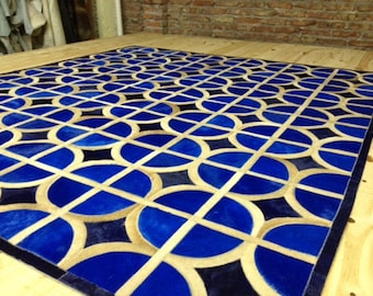 May include: A geometric patterned rug made of leather in shades of blue, tan, and black. The rug features a repeating pattern of circles and squares.