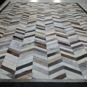 May include: A large rectangular rug with a chevron pattern. The rug is made of various shades of brown, grey, and cream-coloured materials. The edges of the rug are bordered with a black material. The rug is designed for home decor.