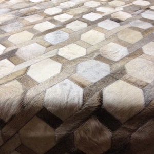 May include: A beige and white cowhide rug with a geometric pattern. The rug is made of individual pieces of cowhide that are sewn together to create a hexagonal pattern.