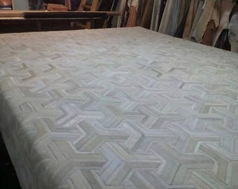 May include: A large rectangular rug featuring a repeating geometric pattern composed of small, interlocking pieces of hide in varying shades of off-white, cream, and light gray, creating a textured surface with a consistent, repeating angular design.