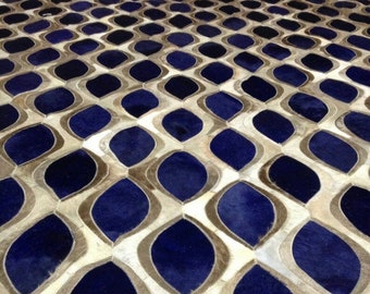 May include: A large area rug constructed from stitched panels of hair-on hide. The surface features a repeating geometric pattern of deep navy blue, almond-toned, and off-white leaf-shaped segments arranged in a continuous, interlocking grid.