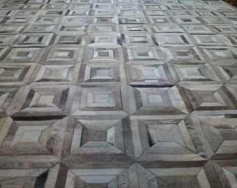 May include: A large area rug constructed from individual square patches of natural cowhide leather. The rug features a repeating geometric pattern of nested squares in varying shades of gray, beige, white, and dark brown, stitched together.