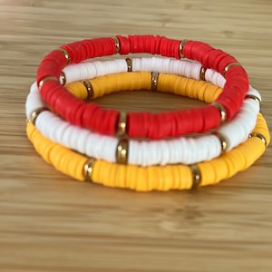 Könnte beinhalten: Drei übereinanderliegende Armbänder mit roten, weißen und gelben Perlen. Jedes Armband hat goldene Akzente.