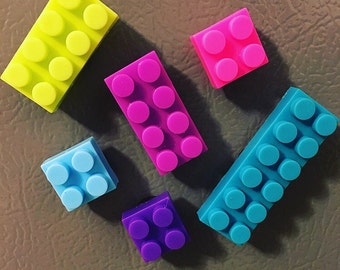 May include: Five colorful plastic toy building blocks in the shape of bricks. The blocks are yellow, pink, purple, blue, and light blue.