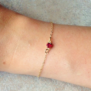 以下が含まれることがあります： 赤い宝石のチャームが1つ付いた、繊細なゴールドチェーンのブレスレットです。