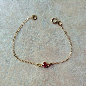 May include: A delicate gold chain bracelet with a single red gemstone.