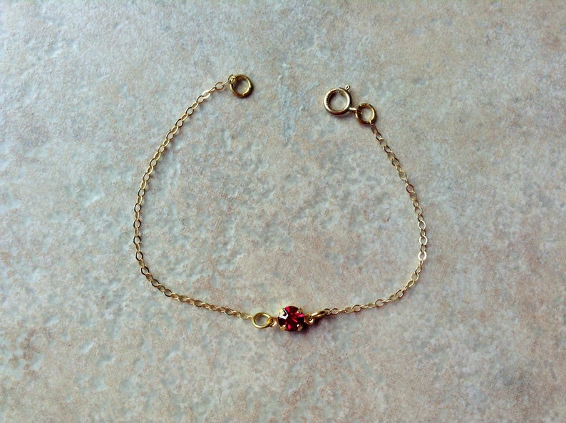 May include: A delicate gold chain bracelet with a single red gemstone.