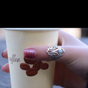 K&ouml;nnte beinhalten: Ein wei&szlig;er Pappbecher mit dem Wort "coffee" darauf. Der Becher wird von einer Hand gehalten, die einen silbernen Ring mit einem blauen Stein tr&auml;gt. Der Ring ist ein Hamsa-Hand-Design.