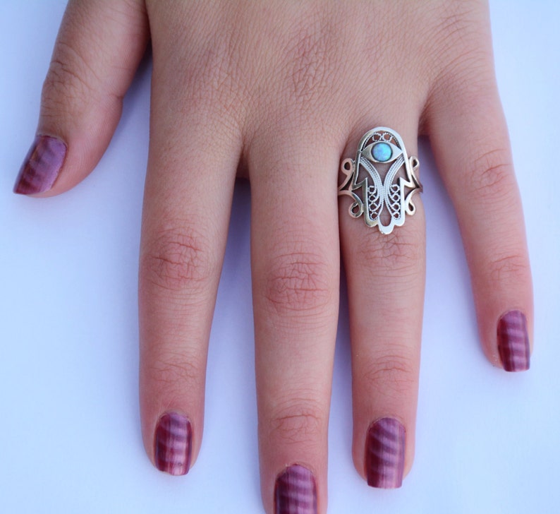K&ouml;nnte beinhalten: Ein silberner Ring mit einem Hamsa-Hand-Design und einem blauen T&uuml;rkis-Stein in der Mitte. Der Ring befindet sich an einem Finger mit burgunderfarbenem Nagellack.
