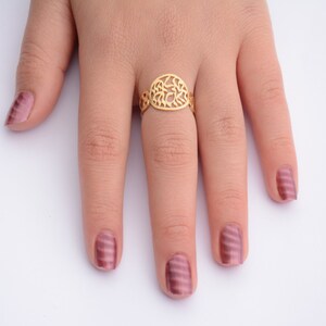 May include: A gold ring with a delicate, intricate design. The ring is worn on the middle finger of a hand with pink nail polish.