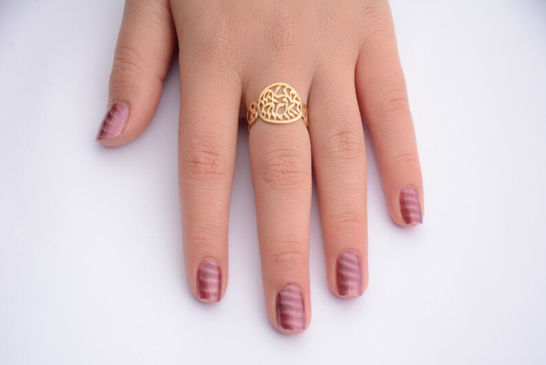 May include: A gold ring with a delicate, intricate design. The ring is worn on the middle finger of a hand with pink nail polish.