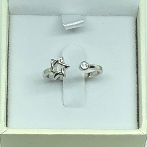 Op de afbeelding: Een zilveren ring met een Davidster-ontwerp aan de ene kant en een enkele heldere strasssteen aan de andere kant. De ring wordt gepresenteerd in een witte doos.