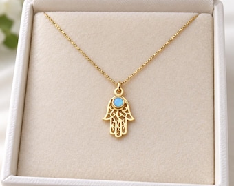 Peut inclure: Un collier doré composé d'une fine chaîne à maillons carrés et d'un pendentif main de Hamsa. Le pendentif présente un motif filigrane complexe avec une petite pierre ronde irisée bleu clair au centre de la paume.