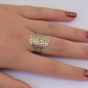 K&ouml;nnte beinhalten: Ein silberner Ring mit einem runden, durchbrochenen Design mit hebr&auml;ischen Buchstaben. Der Ring wird an einem Finger mit rotem Nagellack getragen.