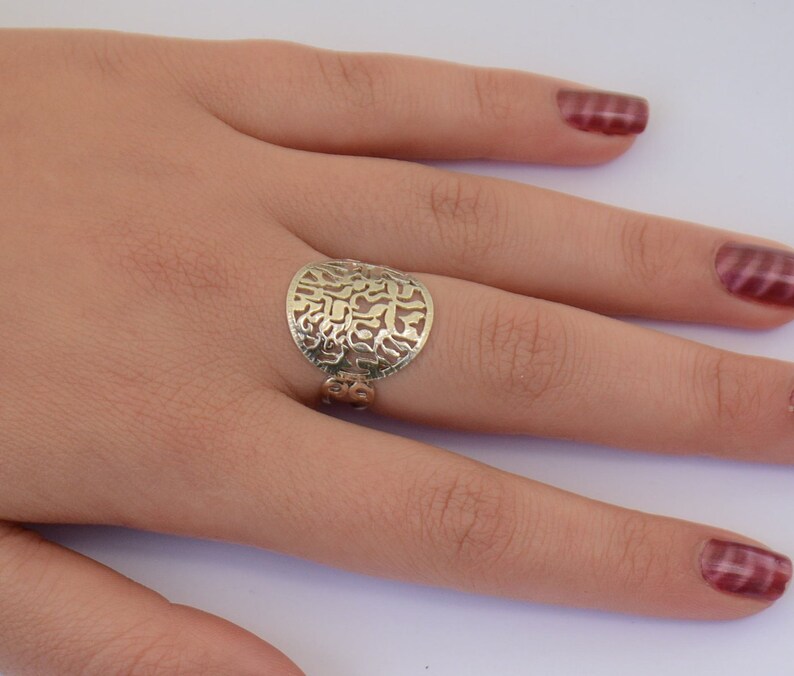 K&ouml;nnte beinhalten: Ein silberner Ring mit einem runden, durchbrochenen Design mit hebr&auml;ischen Buchstaben. Der Ring wird an einem Finger mit rotem Nagellack getragen.