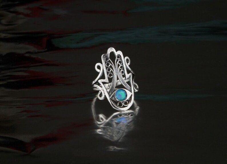 K&ouml;nnte beinhalten: Ein silberner Ring mit einem filigranen Design, das einer Hand mit einem Auge in der Handfl&auml;che &auml;hnelt. Das Auge ist ein blauer Opal-Edelstein.
