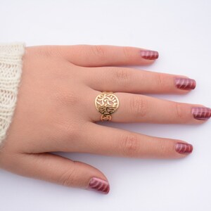 May include: A gold ring with a delicate, circular design featuring a cutout pattern. The ring is worn on the middle finger of a hand with burgundy nail polish.