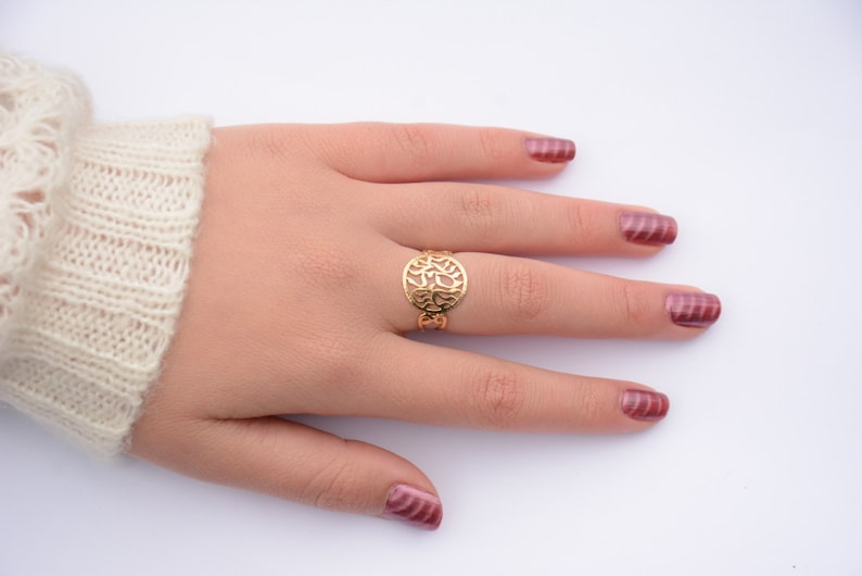 May include: A gold ring with a delicate, circular design featuring a cutout pattern. The ring is worn on the middle finger of a hand with burgundy nail polish.
