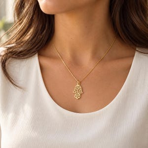 Könnte beinhalten: Eine goldfarbene Halskette mit einer zarten Kette und einem Anhänger in Form einer Hamsa-Hand. Der Anhänger weist ein komplexes, durchbrochenes Filigranmuster auf der gesamten Oberfläche auf und hängt an einer einfachen Gliederkette.