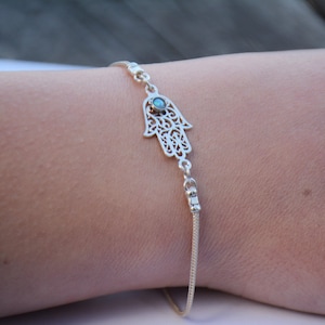 May include: A silver bracelet with a hamsa hand charm featuring a blue opal stone in the center. The hamsa hand is a popular symbol of protection and good luck.