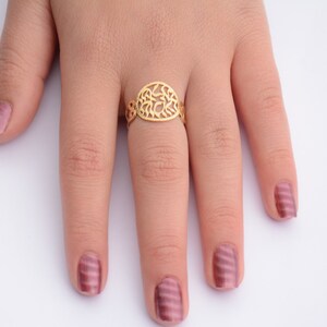 May include: A gold ring with an oval-shaped design featuring intricate cutouts. The ring is worn on a finger with pink nail polish.