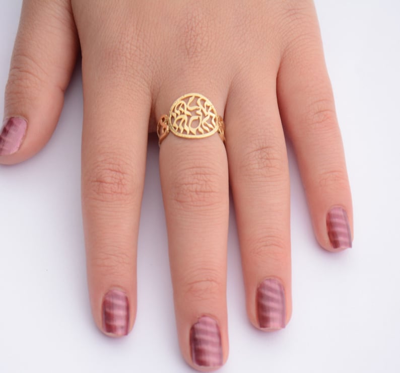 May include: A gold ring with an oval-shaped design featuring intricate cutouts. The ring is worn on a finger with pink nail polish.