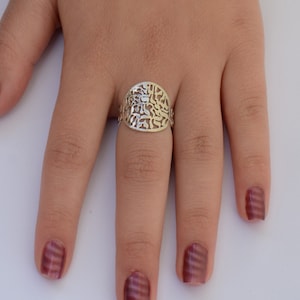 K&ouml;nnte beinhalten: Ein silberner Ring mit einem filigranen Design mit hebr&auml;ischen Buchstaben. Der Ring befindet sich an einem Finger mit burgunderfarbenem Nagellack.