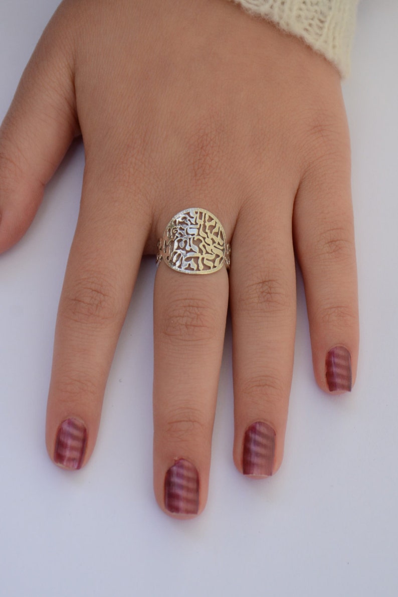 K&ouml;nnte beinhalten: Ein silberner Ring mit einem filigranen Design mit hebr&auml;ischen Buchstaben. Der Ring befindet sich an einem Finger mit burgunderfarbenem Nagellack.