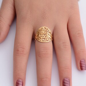 May include: A gold ring with a filigree design featuring Hebrew letters. The ring is worn on a finger with burgundy nail polish.