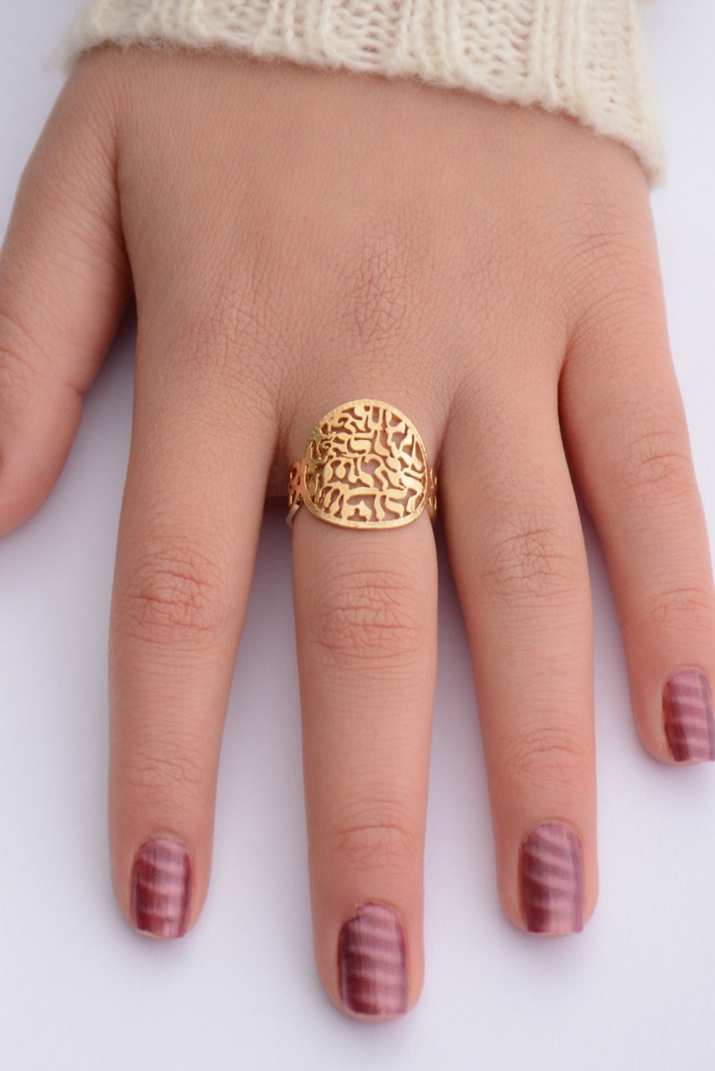 May include: A gold ring with a filigree design featuring Hebrew letters. The ring is worn on a finger with burgundy nail polish.