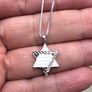 Könnte beinhalten: Eine silberne Kettenkette mit einem silbernen Davidstern-Anhänger. Der Anhänger hat ein strukturiertes, diamantähnliches Muster.