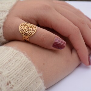 May include: A gold ring with a round, openwork design featuring Hebrew lettering. The ring is worn on a finger with burgundy nail polish.