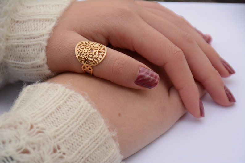 May include: A gold ring with a round, openwork design featuring Hebrew lettering. The ring is worn on a finger with burgundy nail polish.